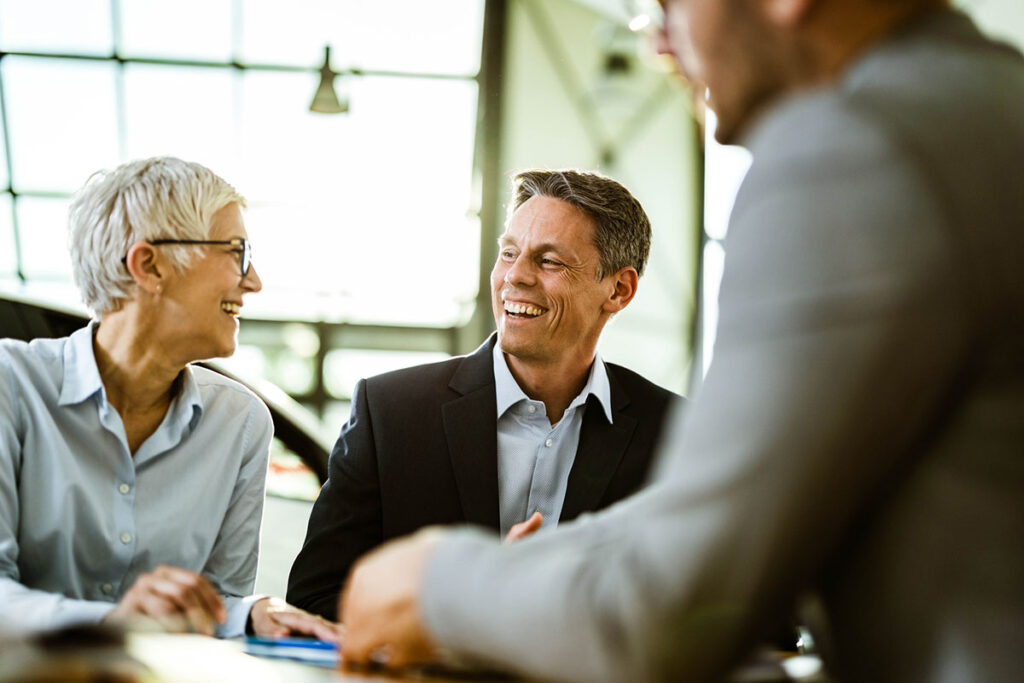 Happy business couple talking to each other while having a meeting with car salesman in a showroom. Focus is on man.