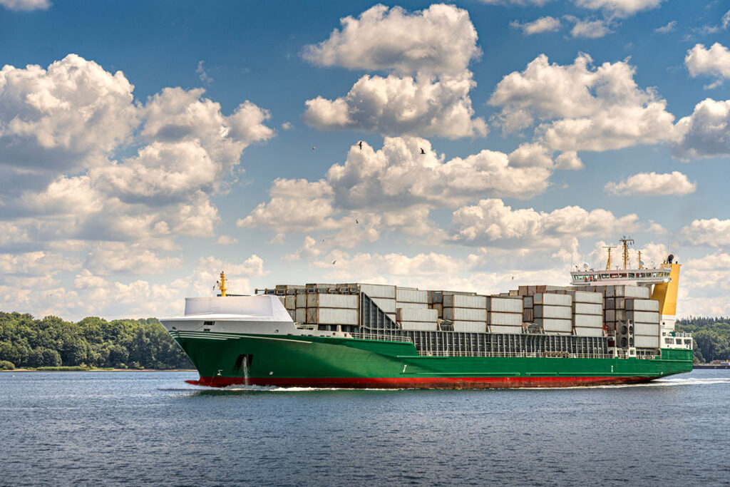 A full loaded cargo ship sets out to sea. Coastal landscape background. Sunny day, blue sky with clouds.