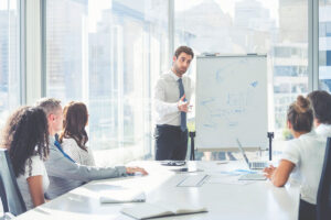 Businessman giving a presentation to his team. He is using a whiteboard with charts and graphs. They are in an office boardroom at the table with laptop computers and paperwork. Men and women in the group.